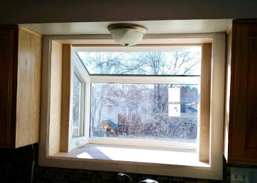Large open casement window framed by wood cabinets, overlooking bare trees and neighboring homes in bright daylight.