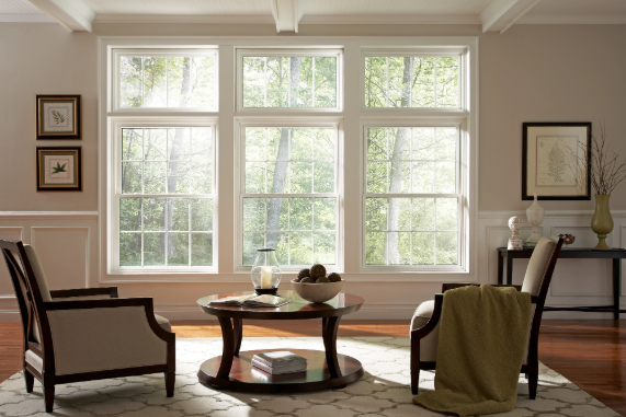 Bright living room with three large white-framed windows, two armchairs, and a round table overlooking leafy trees.